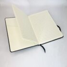 An open, black hardcover notebook with cream-colored, lined pages on a white surface. A black ribbon bookmark is visible, and the notebooks back cover is partially open.