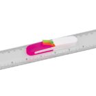 A transparent plastic ruler showing both metric and imperial measurements. It has a pink and white pen-shaped holder attached at the center, containing small, colorful page flags.
