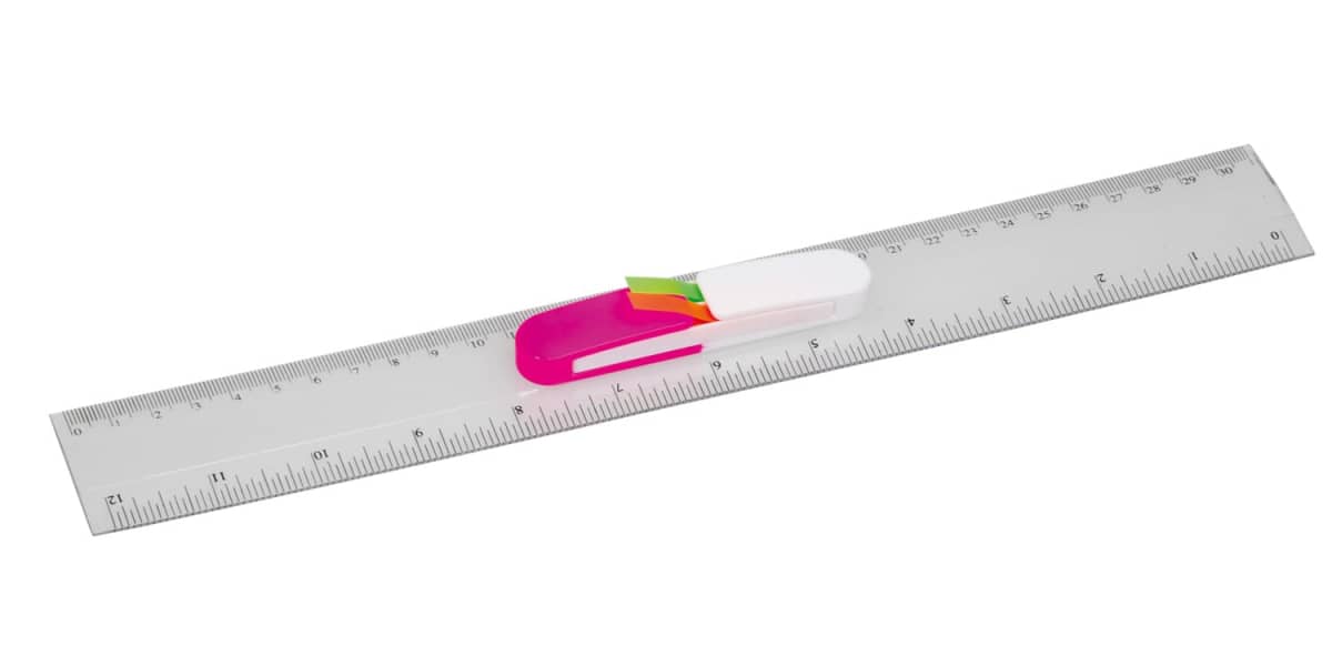A transparent plastic ruler showing both metric and imperial measurements. It has a pink and white pen-shaped holder attached at the center, containing small, colorful page flags.