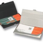 Two business card holders: one black with a closed lid, the other silver with an open lid displaying a card. The cards have a modern design with orange, green, and gray colors.