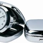 A small, silver-colored portable travel clock with a white face and black numbers, set in a shiny, clamshell case. One clock is open showing the time, while another is closed, highlighting the metallic exterior.