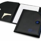 A black faux leather portfolio folder is open to reveal a lined notebook inside. The left flap contains a pocket with sticky notes. The folder has a button clasp and features a sleek, modern design.