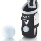 A small black golf accessory pouch with a carabiner clip, labeled APC, holds a white ball marker, a divot tool, and three golf tees. A white golf ball is positioned next to the pouch.