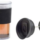 A transparent travel mug with a black grip and a removable lid is shown. Next to it is a black plunger with a mesh filter, designed for making coffee or tea on the go. The mug contains a brown liquid and some coffee grounds at the bottom.