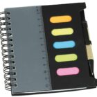 A small spiral notebook with a ruler along the left edge. It features four colorful sticky note pads in orange, blue, green, and pink, and has a pen attached to the side.