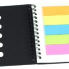 An open spiral-bound notebook with a black cover showing colorful sticky notes in orange, yellow, blue, green, and pink on the right page. A black pen is clipped to the notebooks side.