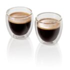 Two double-walled glass cups filled with rich espresso, displaying a light crema on top, are placed on a reflective white surface.