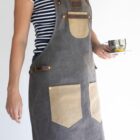 Person wearing a striped shirt and a gray apron with tan pockets and leather straps, holding a clear cup with a lemon wedge on a saucer. The apron has several buttons and a rectangular patch on the chest. The background is plain white.