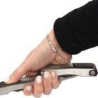 A hand holding a multi-tool with a silver and black handle, featuring an open bottle opener. The person is wearing a silver bracelet and a black sleeve.
