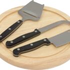 A round wooden cheese board with three cheese tools: a slicer, a fork-tipped knife, and a mini cleaver. All have black handles with silver rivets.