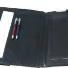 A black leather portfolio with a snap closure lies open. It contains a notepad with a logo, two pens (one red and one blue) in pen slots, and a narrow pocket. A silver and black pen is clipped to the top edge.