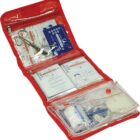 A red first aid kit is open, displaying various medical supplies such as bandages, gauze, gloves, and informational pamphlets. The kit is organized in clear plastic compartments with items neatly arranged.