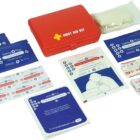 A red first aid kit is open, displaying its contents. Items include antiseptic wipes, adhesive bandages, gauze pads, and a first aid manual. The kit has a white cross and First Aid Kit label on the lid.