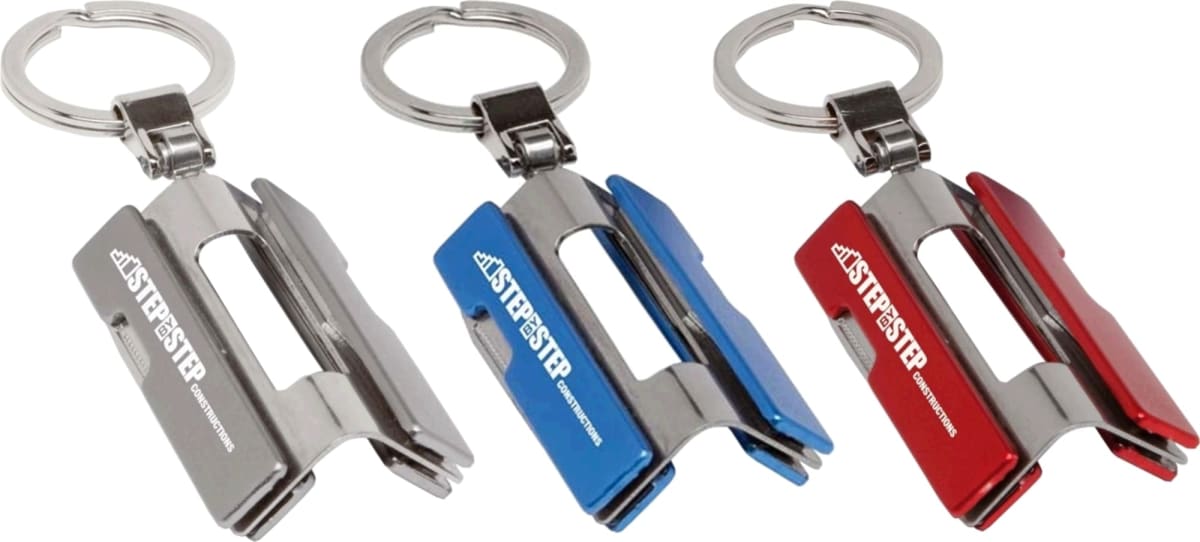 Three metallic keychains, each shaped like a miniature stapler, are displayed. They come in silver, blue, and red, with the word STAP-STEP printed on each. All have a ring attachment for holding keys.