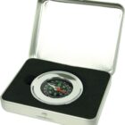 A round compass with a black face and a silver rim sits inside a rectangular metal tin box. The box has dark foam padding with a circular cutout to hold the compass securely.