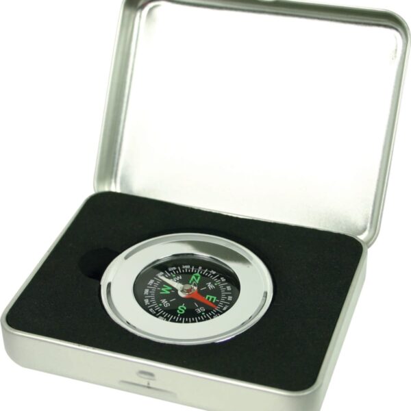 A silver compass with a black and green dial is placed inside a rectangular metal box with a black foam interior. The box lid is partially open, showing the compass clearly.