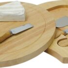 Round wooden cheese board with a slide-out drawer containing three cheese knives and two wedges of white cheese on top.