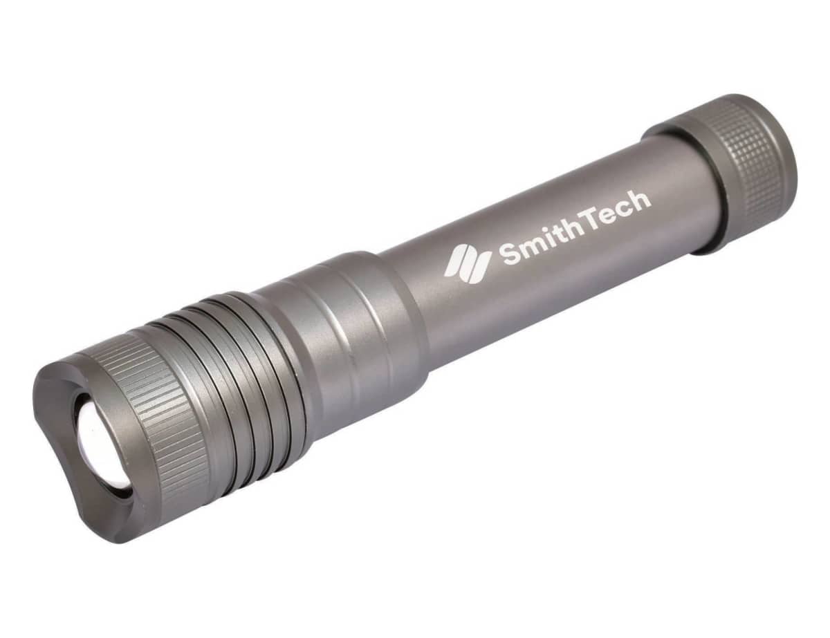 A silver flashlight with a textured grip and the brand name SmithTech printed on the side. The flashlight has a cylindrical shape with a flat, rounded end.