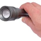 A hand holding a small black flashlight with a textured grip and a lens on the front, against a plain white background.