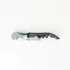 A metal corkscrew with an attached bottle opener and serrated foil cutter rests against a white background. The handle is black, and the screw is extended, ready for use.