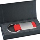 A keychain with a rectangular metal tag and a red strap is displayed in a black box with a foam lining.