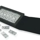 A black wristwatch designed to resemble a domino set, with a rectangular face showing domino dots. Several small dominos are scattered alongside the unbuckled black strap.