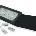 A set of white dominoes with black dots, placed on a black surface. Some dominoes are laid out, while others are arranged in a rectangular tray with a lid. The setup appears to be designed for travel or compact storage.