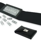 A set of white dominoes with black dots displayed next to an open black leather case designed to hold them. The case has a secure buckle and lies flat, showing its interior and a few dominoes scattered around it.