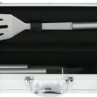 A silver metal case is open to reveal grilling tools inside, including a spatula. The tools are secured in a black foam insert. The case has clasps and a handle for carrying.