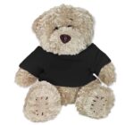 A fluffy beige teddy bear wearing a black T-shirt is sitting. The bear has stitched patches on its paws and a dark brown nose, and it is positioned against a plain white background.