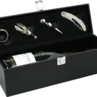 A black wine gift box containing a wine bottle and accessories: a corkscrew, a bottle stopper, a wine pourer, and a drip ring. The box has a metal latch and organized compartments for each item.
