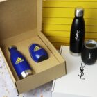 Open cardboard box with two blue Adidas thermoses next to two black thermoses with white YSL logos. One YSL bottle is on top of a white box. Background features a yellow panel and a terrazzo surface.
