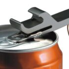 A silver can opener keychain is being used to open an orange soda can. The opener is positioned under the tab of the can, demonstrating its functionality. The keychain ring is visible at the end of the opener.