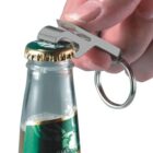 Close-up of a hand using a silver bottle opener attached to a keychain to remove the cap from a beer bottle. The bottle has a green label, and some foam is visible near the top.