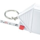 A small, white rectangular tape measure keychain with a retractable tape, featuring a red button. The tape is partially extended, displaying metric and imperial measurements.