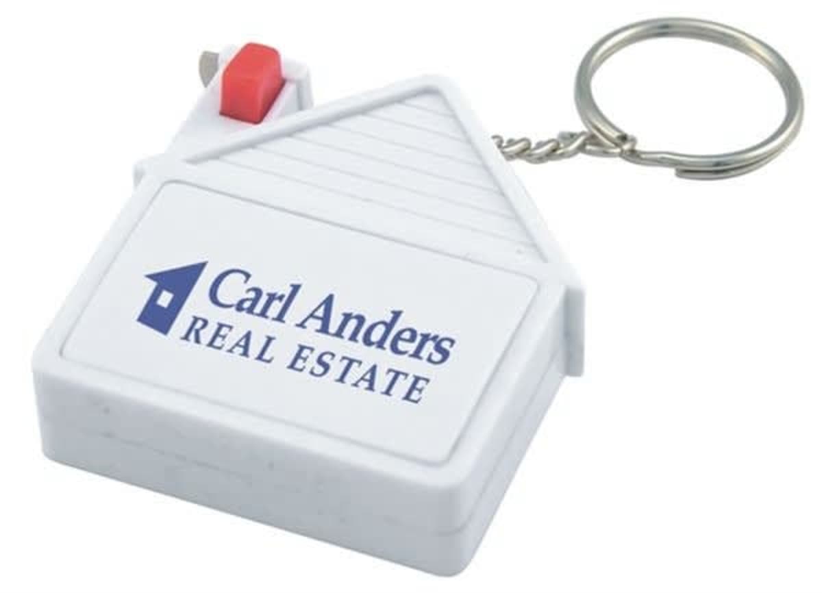 A keychain shaped like a small white house with a red button and a metal key ring attached. It features the logo and text Carl Anders Real Estate in blue on the side.