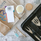 A jute tote bag, custom-branded notepad, scented candle with lid, small glass bottle with mint label, pen, and a black case with barbecue tools are laid out on a speckled surface.