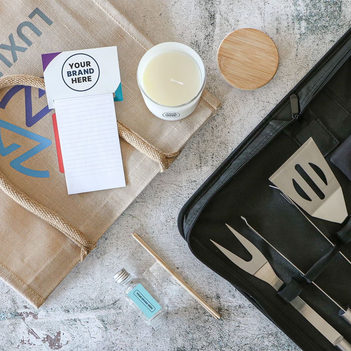 A jute tote bag, custom-branded notepad, scented candle with lid, small glass bottle with mint label, pen, and a black case with barbecue tools are laid out on a speckled surface.
