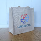 A beige tote bag with the word LUXURIOUS and a colorful geometric design printed on it. The bag is placed on a wooden floor against a white paneled wall.