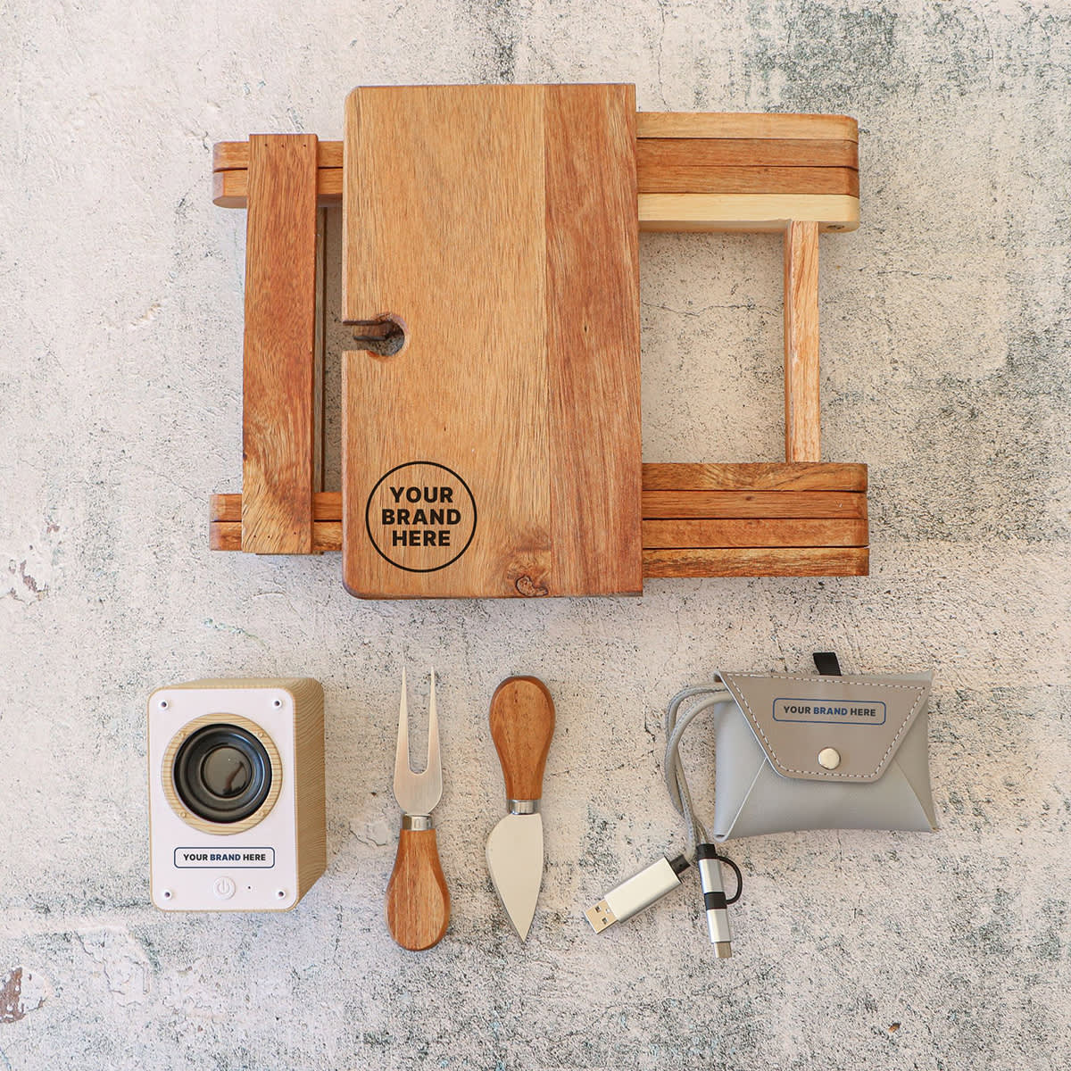 A flat lay image featuring a wooden cheese board set with logo space, a camera, two cheese knives, a cheese fork, and a gray pouch containing a USB cable, all on a textured stone background.