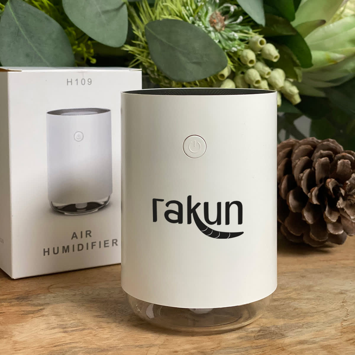 A white Rakun air humidifier with a power button on its front is placed on a wooden surface. It is next to its packaging box, with green foliage and a pine cone in the background.
