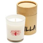 A white candle in a clear glass holder with a red Stuckey Classic Tyres logo, featuring a winged emblem. It is next to a cylindrical brown cardboard container with Vanilla partially visible on the front.