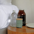 A wooden bedside table holds a clear glass water bottle, a brown jar labeled Mayde Tea, and a green candle with a wooden lid labeled Bask & Stow. The edge of a white bed and a white plate are visible.