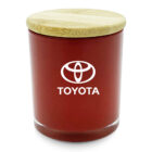 A red candle in a glass jar with a light wooden lid. The jar features the Toyota logo and name in white on the front.