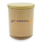 A beige cylindrical candle with a smooth wooden lid. The front of the candle displays the Jet Airways logo in dark blue, with a stylized yellow emblem resembling a wing.