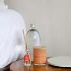 A bedside table with a small glass bottle of reed diffuser, a clear bottle of water, and a red Bask & Stow candle with a wooden lid. A portion of a white bed is visible on the left.