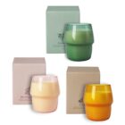 Three colorful candles in glass holders: green, yellow, and peach, each with its own matching box. The boxes feature decorative logos and text.