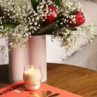 A vase with red flowers and babys breath sits on a wooden table next to a lit LOréal candle and an orange book. The setting creates a cozy and inviting atmosphere.