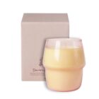 A cream-colored, textured candle in a transparent glass holder is placed in front of a beige box with minimalist design and text.