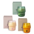 Three candles in unique glass containers with different logos: top green with Shopify, bottom left cream with a pink ribbon, bottom right orange with a logo. Each has a matching rectangular box behind them.